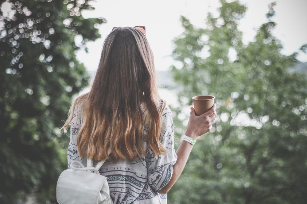 estudiante con pelo largo rubio de espaldas con café y mochila mirando la naturaleza de Canadá